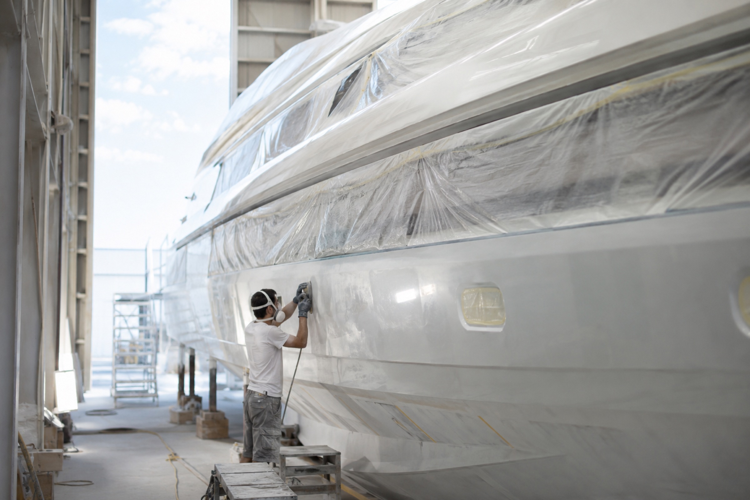 A refit yard worker fairing a yacht hull under cover